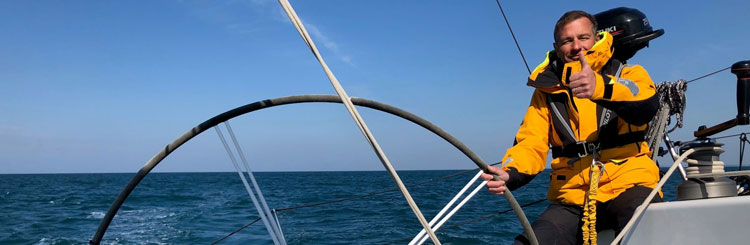 Photo aboard a sailboat of a person in an orange vest holding the helm looking at the lens with a raised thumb